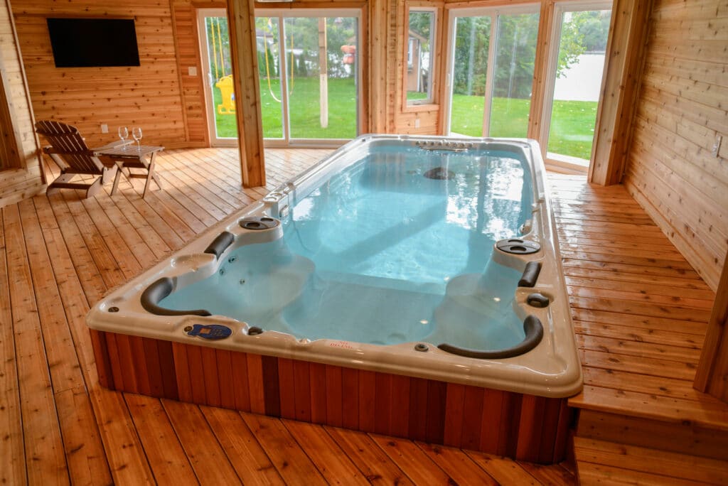 Indoor swim spa with a stone exterior, surrounded by a wooden deck and several lounge chairs, and a scenic view of trees and mountains in the background